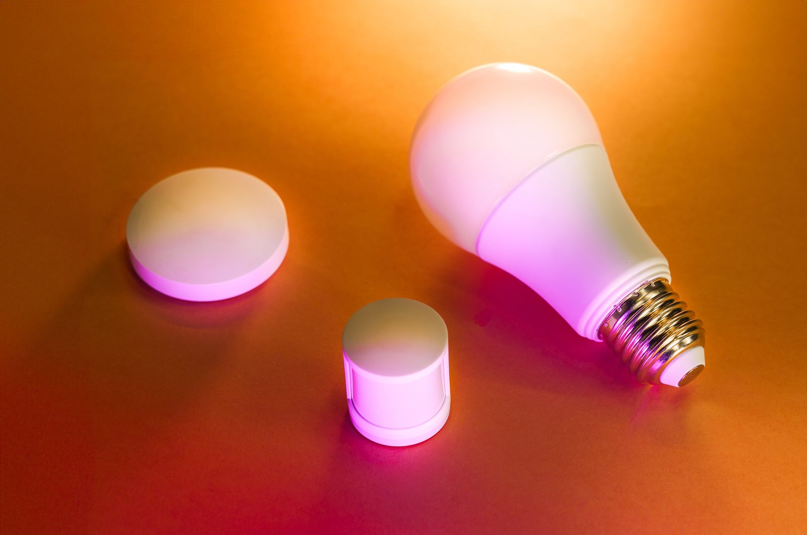 A smart bulb and two small smart-home sensors lit in pink and orange